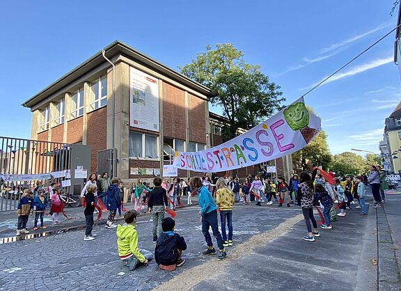 Kinder verschiedenen Alters spielen und bewegen sich auf einer gepflasterten Straße vor einem zweistöckigen Backsteingebäude. Über der Straße hängt ein buntes Banner mit der Aufschrift „Schulstraße“ und einem grünen Baum-Symbol. Im Hintergrund sind Erwachsene zu sehen, die die Aktion begleiten.