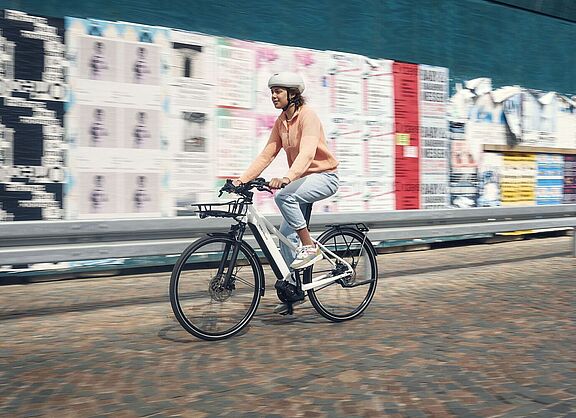 Eine Frau fähet auf einem weißen Elektrorad vor einer Wand mit vielen Plakaten. 